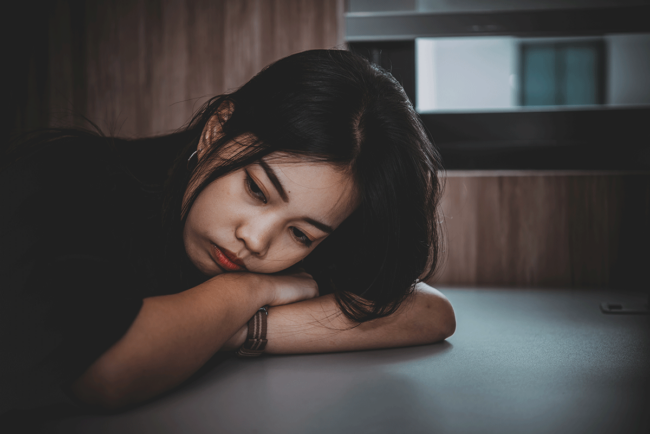 How-to-Recognize-the-Signs-You-Need-Opioid-Treatment a young woman rests her head on her arms on a table in the dark struggling with the realization she might need opioid treatment
