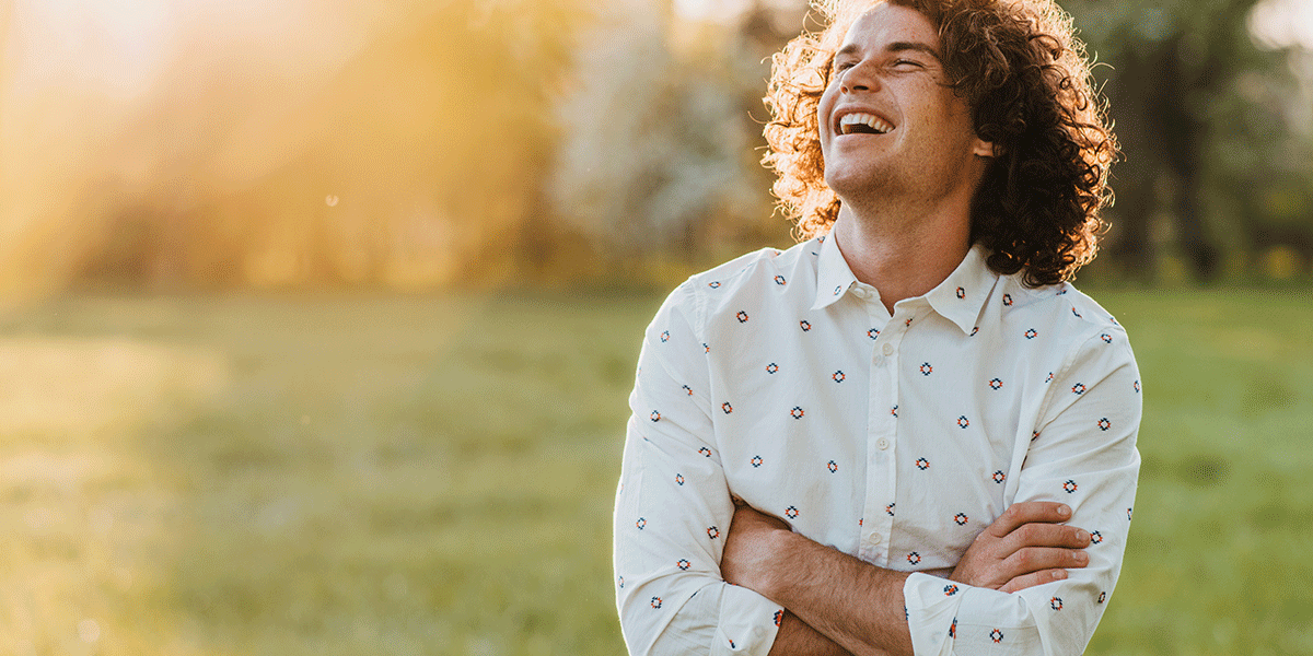 10-Tips-to-Stay-Sober-This-Summer curly haired man with arms crossed stands outside and looks up at the sky while thinking about tips to stay sober