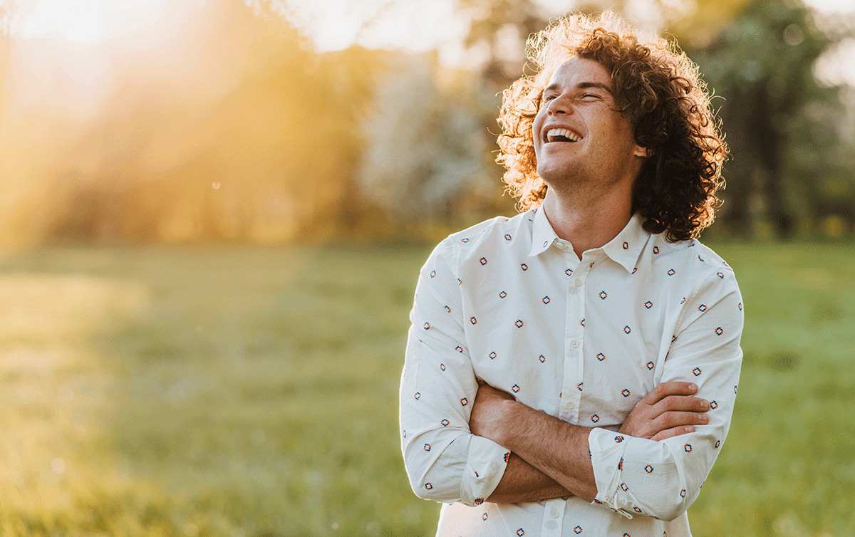 10-Tips-to-Stay-Sober-This-Summer curly haired man with arms crossed stands outside and looks up at the sky while thinking about tips to stay sober