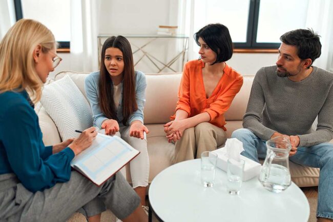 a family of three sits on a couch discussing with their therapist about techniques used in family therapy and the therapist takes notes in her notebook