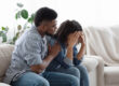 a man hugs his wife while they sits on the couch in distress and struggling with the reality of being married to an alcoholic
