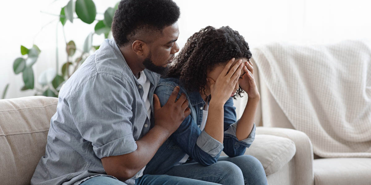 a man hugs his wife while they sits on the couch in distress and struggling with the reality of being married to an alcoholic