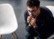 man wearing glasses sits in a chair and leans over thinking about the symptoms of adderall abuse