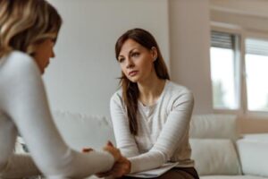 a woman with long hair sits on a couch and holds the hands of a woman sitting across from her while telling her the benefits of cognitive-behavioral therapy