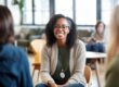 a woman wearing glasses sits with other people in a php drug rehab and attend a group therapy session