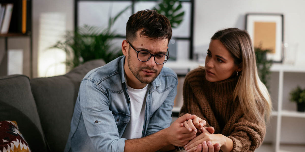How to Help an Alcoholic Who Doesn’t Want Help a man with glasses sits with a woman on a couch as they hold hands and talk about how to help an alcoholic who doesn't want help