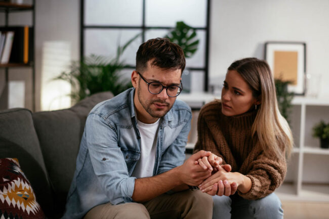 How to Help an Alcoholic Who Doesn’t Want Help a man with glasses sits with a woman on a couch as they hold hands and talk about how to help an alcoholic who doesn't want help