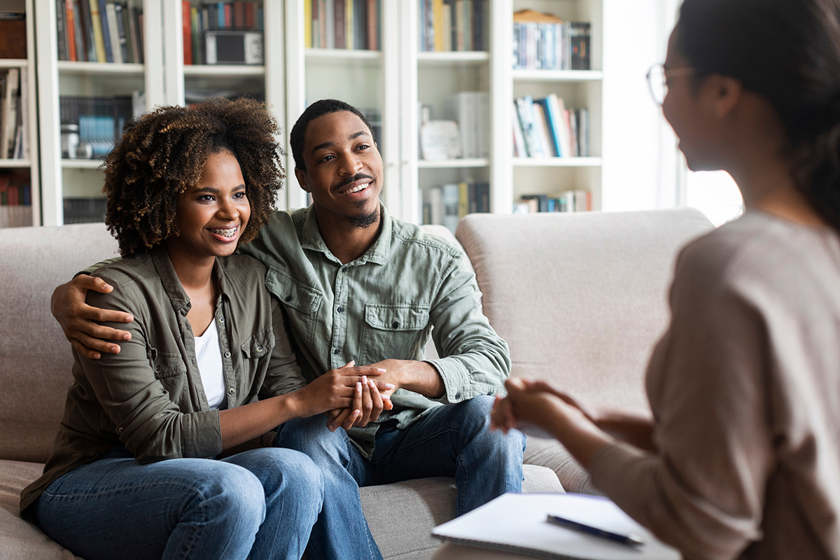 a couple sits on a couch hugging each other talking to therapist explain the benefits of family therapy for substance abuse recovery
