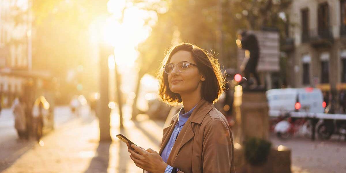 Navigating-Life-After-Rehab-A-Step-by-Step-Guide smiling woman with glasses holds her phone while outside enjoying her life after rehab