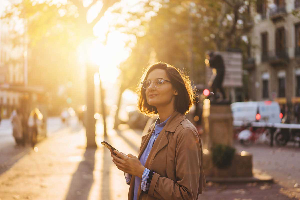 Navigating-Life-After-Rehab-A-Step-by-Step-Guide smiling woman with glasses holds her phone while outside enjoying her life after rehab