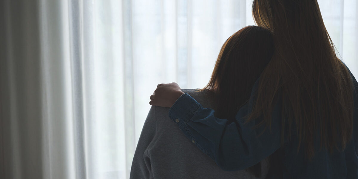 Tips to Help a Loved One in Recovery two people hug while facing a curtained window and talking about the importance of helping a loved one in recovery