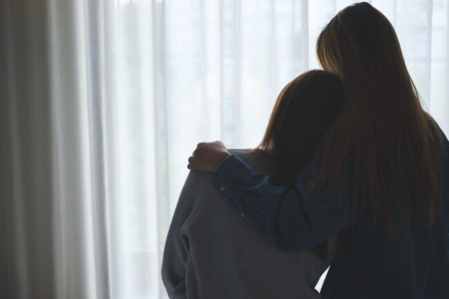 Tips to Help a Loved One in Recovery two people hug while facing a curtained window and talking about the importance of helping a loved one in recovery
