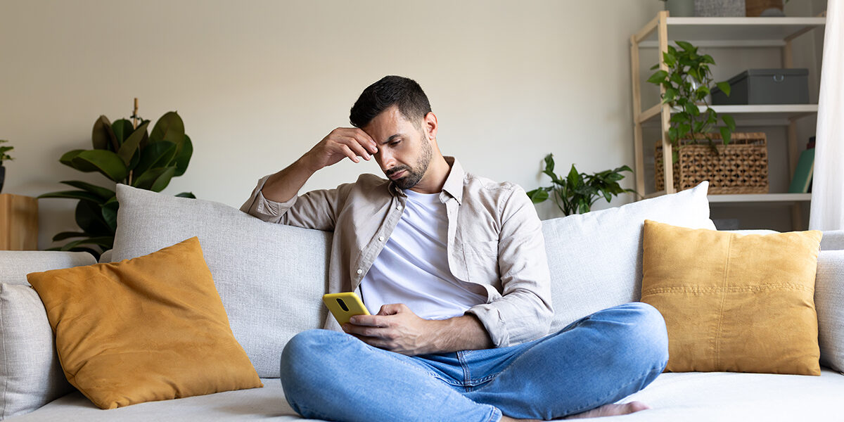 The-Impact-of-Social-Media-on-Addiction-Recovery a distraught man sits on his couch and holds his hand to his forehead in distress about the impact of social media on his. addiction recovery