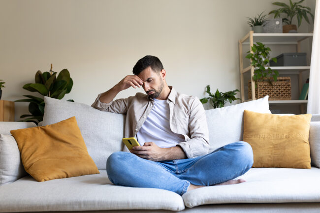 The-Impact-of-Social-Media-on-Addiction-Recovery a distraught man sits on his couch and holds his hand to his forehead in distress about the impact of social media on his. addiction recovery