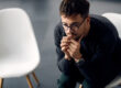 man with glasses sits in a chair next to an empty chair and thinks about the link between trauma and addiction