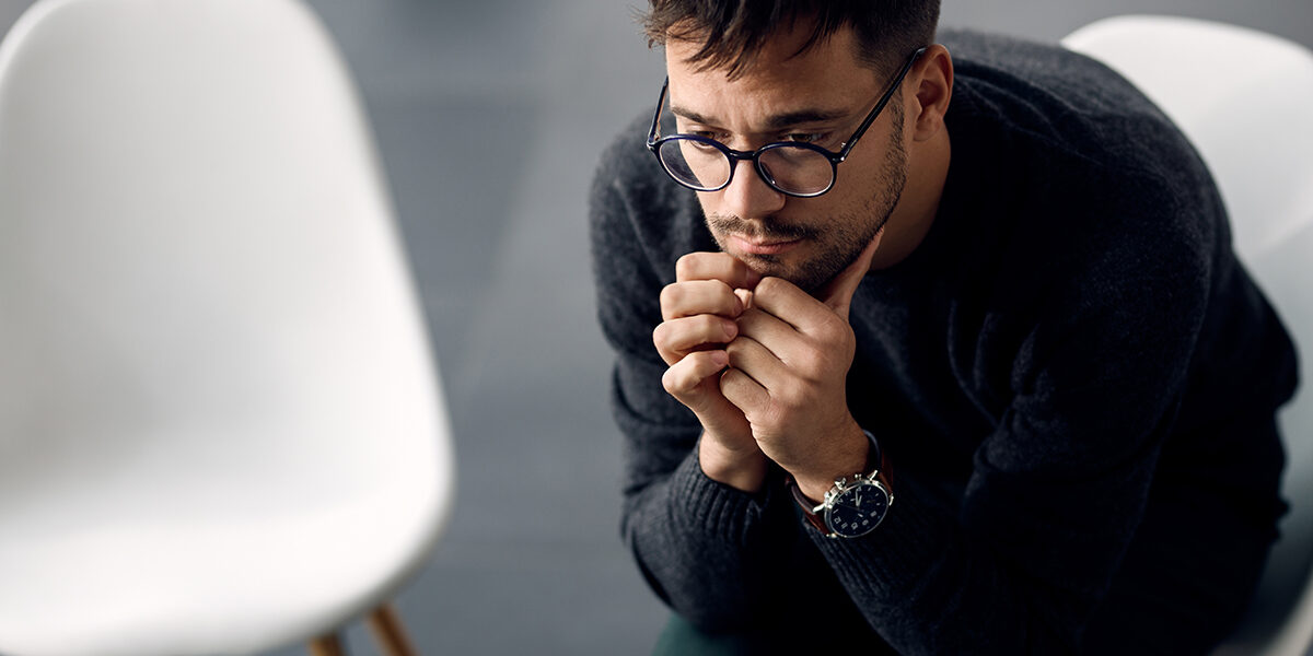 man with glasses sits in a chair next to an empty chair and thinks about the link between trauma and addiction