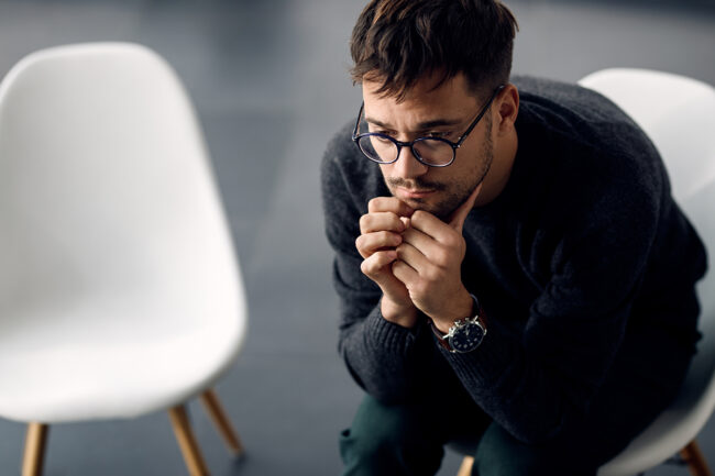 man with glasses sits in a chair next to an empty chair and thinks about the link between trauma and addiction