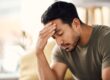 Man sitting indoors with his hand on his forehead and eyes closed, appearing stressed or deep in thought, suggesting feelings of worry, fatigue, or emotional strain.