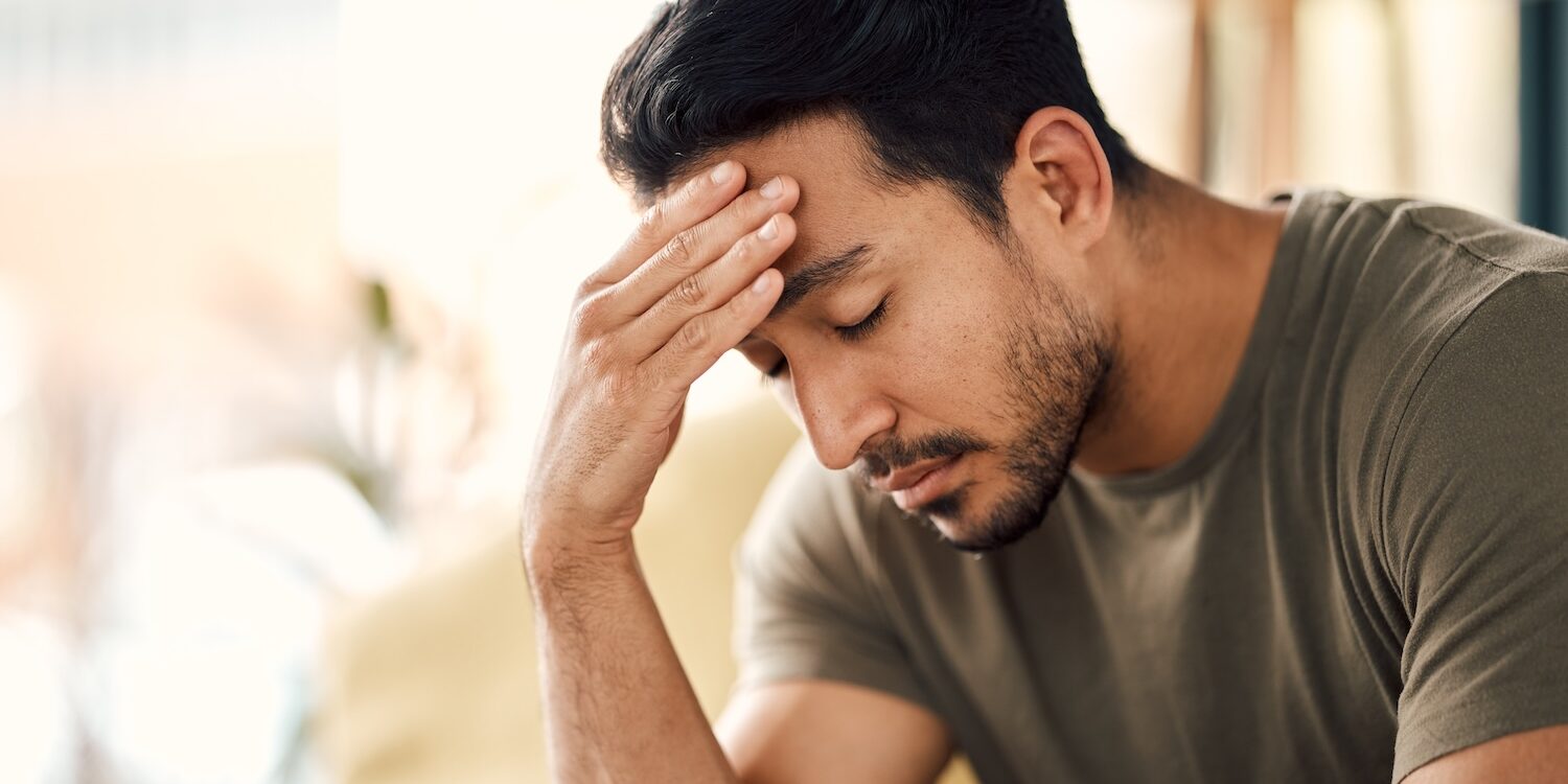 Man sitting indoors with his hand on his forehead and eyes closed, appearing stressed or deep in thought, suggesting feelings of worry, fatigue, or emotional strain.