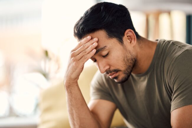 Man sitting indoors with his hand on his forehead and eyes closed, appearing stressed or deep in thought, suggesting feelings of worry, fatigue, or emotional strain.