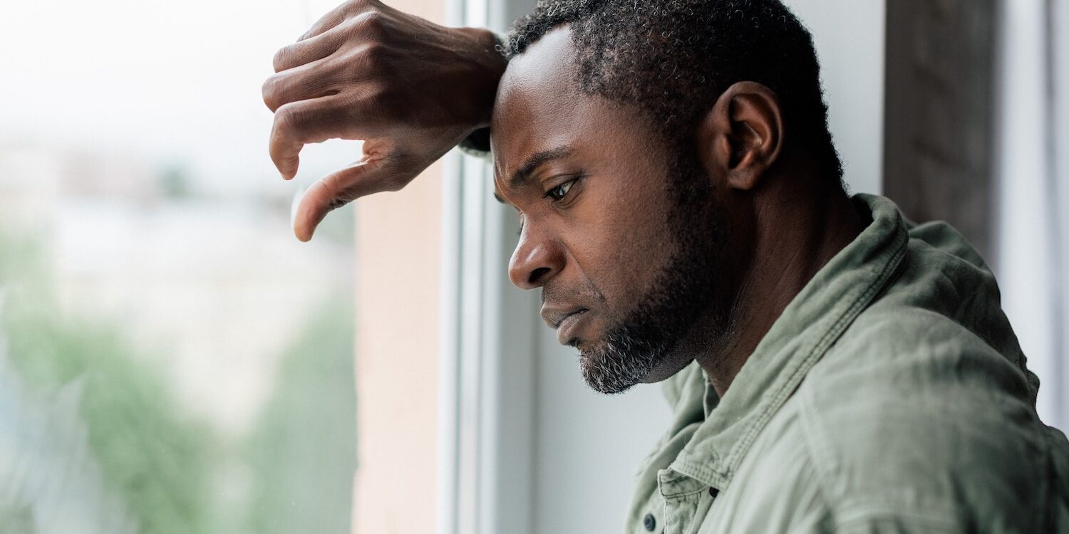 Man standing by a window with his arm resting on the frame, gazing downwards with a pensive, somber expression, suggesting feelings of sadness, contemplation, or fatigue.