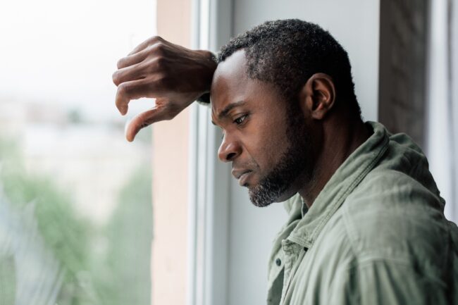 Medically Managed Withdrawal at Spokane Falls Recovery Center Man standing by a window with his arm resting on the frame, gazing downwards with a pensive, somber expression, suggesting feelings of sadness, contemplation, or fatigue.