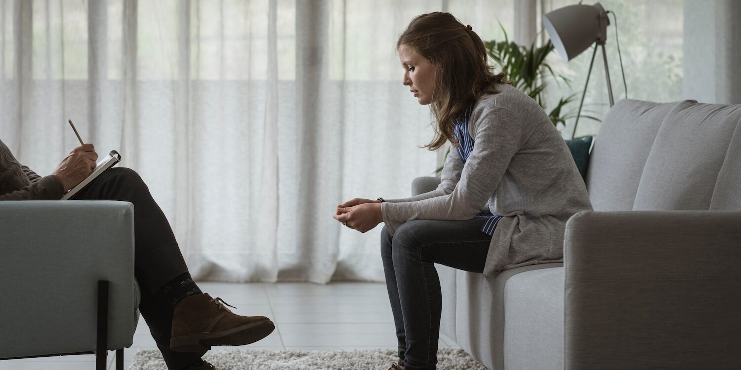 Woman sitting on a couch leaning forward with a thoughtful, tense expression during a therapy session, while a therapist across from her takes notes in a calm, softly lit room.