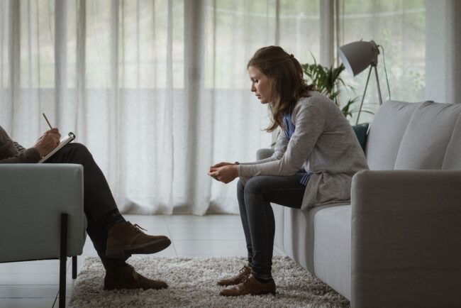 Woman sitting on a couch leaning forward with a thoughtful, tense expression during a therapy session, while a therapist across from her takes notes in a calm, softly lit room.