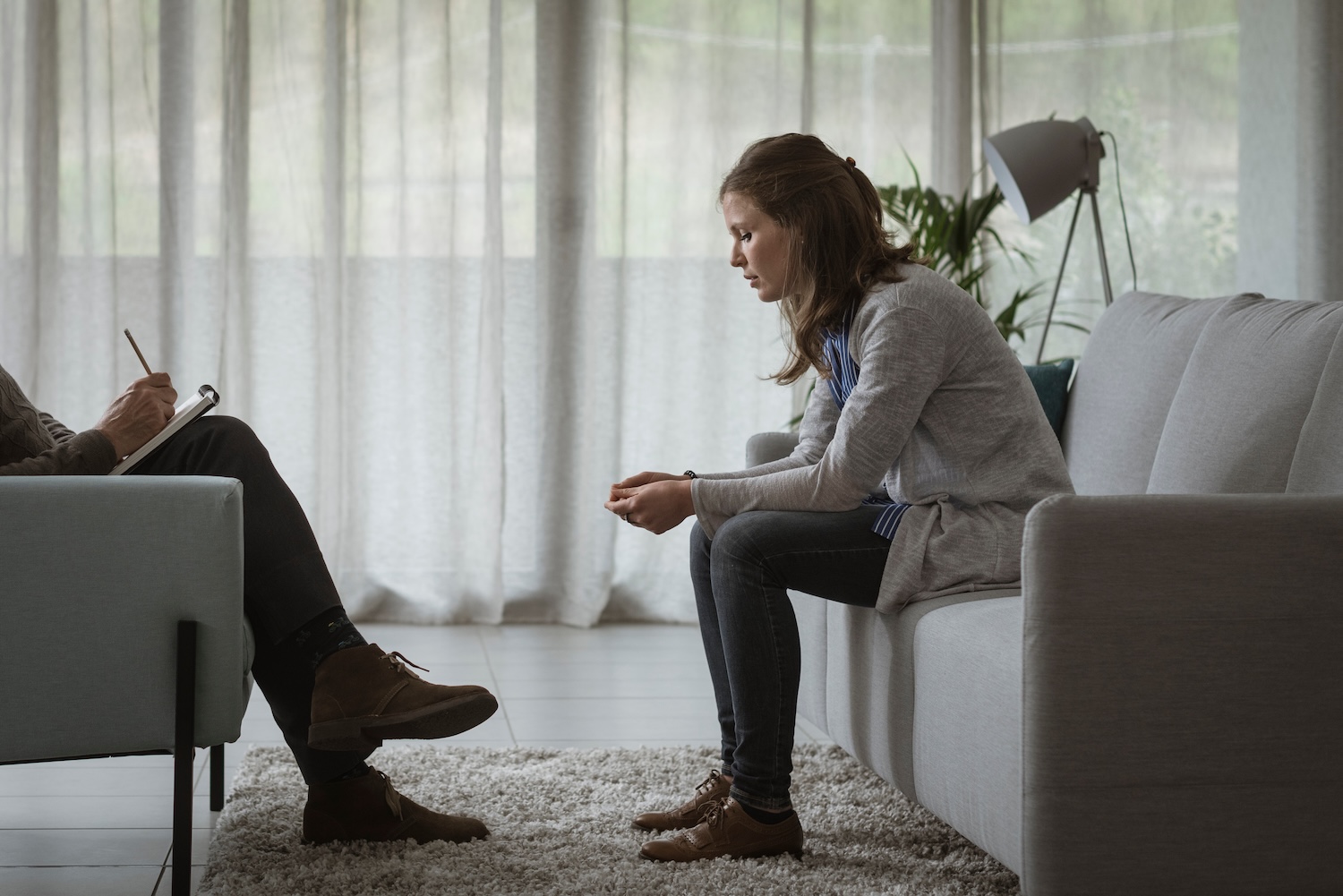 Therapist listening to the patient during a psychotherapy session Woman sitting on a couch leaning forward with a thoughtful, tense expression during a therapy session, while a therapist across from her takes notes in a calm, softly lit room.