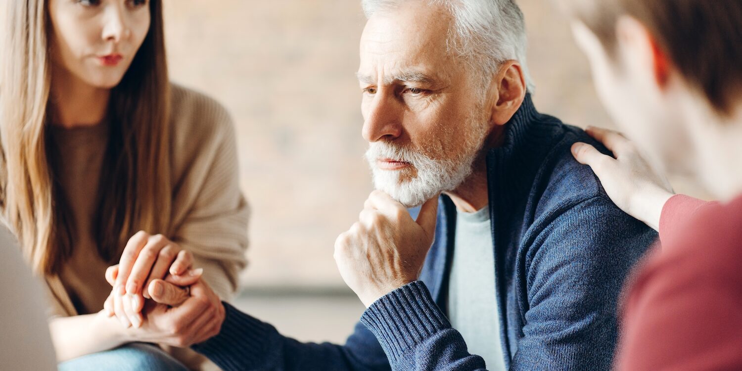 Elderly man sitting with a thoughtful, distressed expression while two younger people sit close, holding his hand and resting a hand on his shoulder in a gesture of empathy and support.